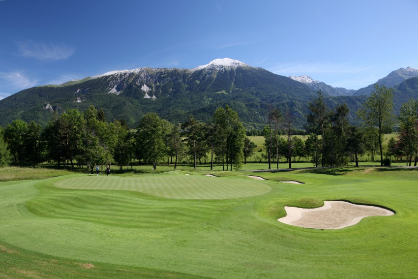 Golf igrišče s peščenimi ovirami, obdano z drevesi in s pogledom na hribe.